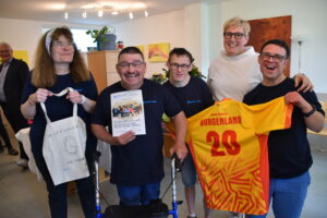 Drei Bewohner und eine Bewohnerin der Wohngemeinschaft halten stolz ein Trikot der special Olympics mit der Aufschrift "Burgenland 20", die Einladung zum gemeinsamen Fest und eine selbst gestaltete Stofftasche in die Kamera.