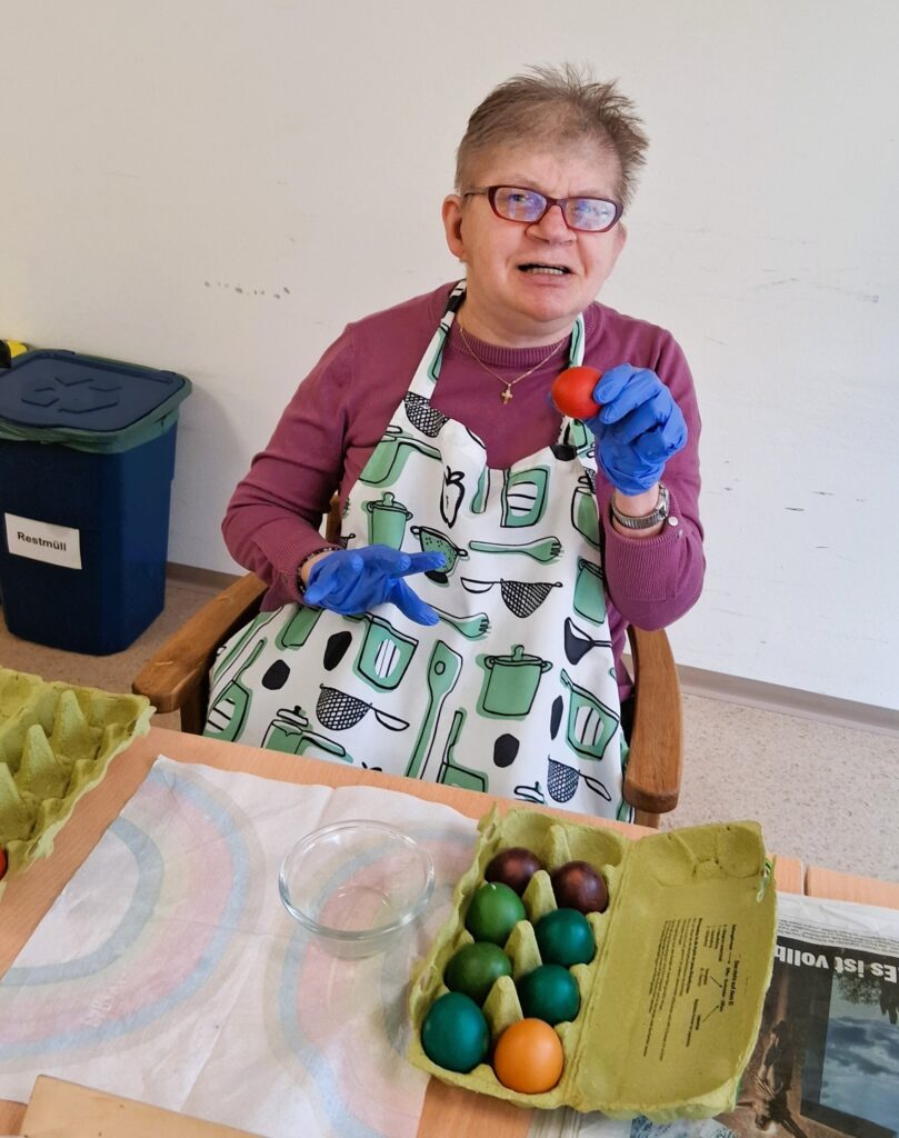 Eine Klientdin der betreuten Wohngemeinschaft Jennersdorf trägt blaue Gummihandschuhe und eine Schürze. Sie hält ein gefärbtes Ei in die Kamera. Vor ihr auf dem Tisch liegen noch mehr gefärbte Eier in einem Eierkarton.