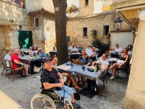 In einem schattigen Innenhof haben es sich die Bewohner:innen der betreuten Wohngemeinschaft Eisenstadt an mehreren Tischen bequem gemacht. Alle blicken lachend in die Kamera.