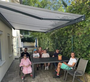 Bewohner:innen der betreuten Wohngemeinschaft Neudörfl haben es sich im Schatten rund um einen großen grauen Alu-Tisch gemütlich gemacht.