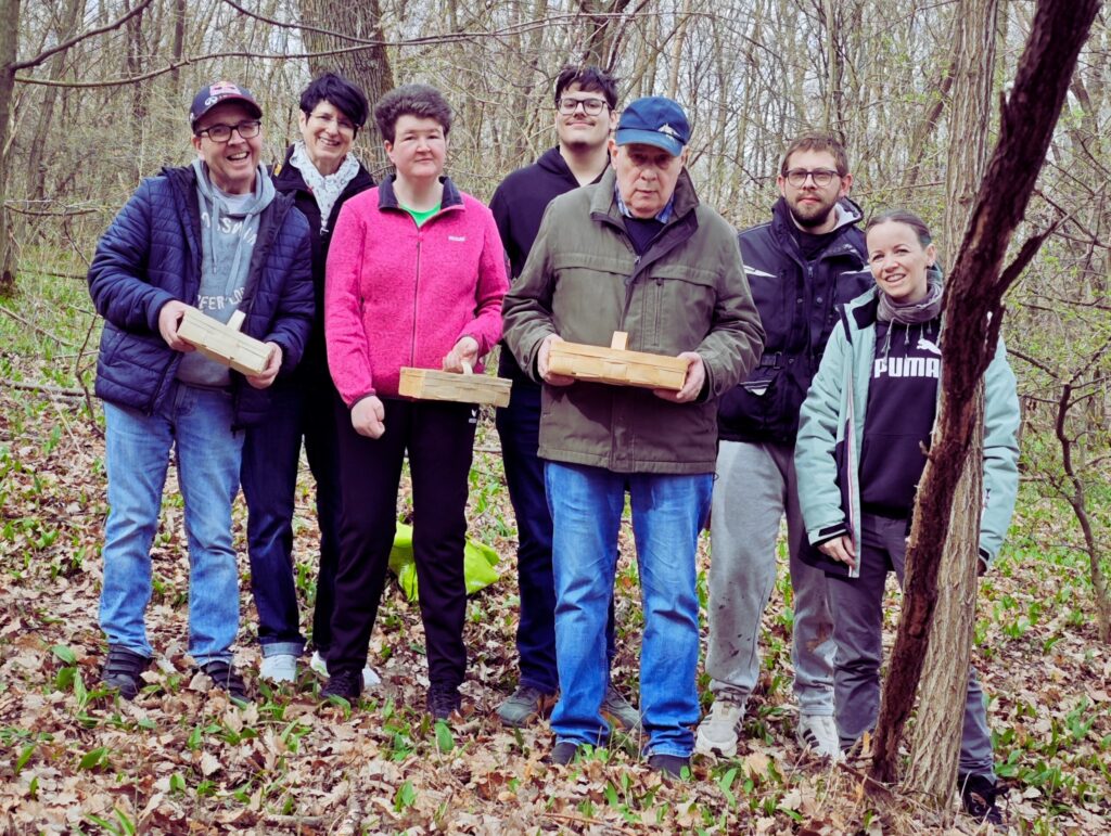 Die KlientInnen und MitarbeiterInnen der Förderwerkstätte Oberpullendorf lächeln in die Kamera. Sie stehen im Wald und einige halten Körbchen mit selbst gesammelten Bärlauch in der Hand.