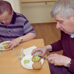 Zwei Klienten sitzen gemeinsam an einem Tisch. Beide haben ein Teller vor sich und streichen den selbstgemachten Bärlauchaufstrich auf ein Brot.