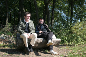 Walter Paulhart sitzt mit einem zweiten Mann auf einer aus einem Holzstamm gefertigten Sitzbank auf einer Waldlichtung. Die Männer unterhalten sich lachend.