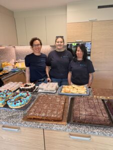 Drei Betreuerinnen posieren stolz vor dem vorbereiteten Kuchenbuffet.