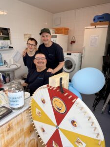 Zwei Bewohner posieren gemeinsam mit einem Betreuer. Vor ihnen steht ein Glücksrad und ein Sammelglas für Spenden.