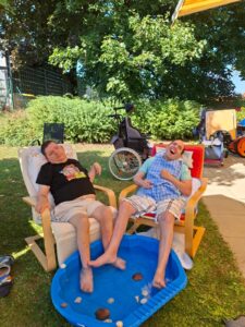 Im Garten des Tageswohnheims St. Margarethen wurde ein kleines blaues Plantschbecken aufgestellt. Zwei Bewohner sitzen in Sesseln um das Plantschbecken und kühlen ihre Füße im kalten Wasser.