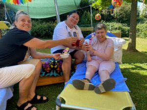Im Garten des Tageswohnheims St. Margarethen sitzt eine Bewohnerin auf einem gemütlichen Liegestuhl. In der Hand hält sie ein Glas mit rotem Fruchtsaft und prostet zwei neben ihr sitzenden Betreuerinnen zu.