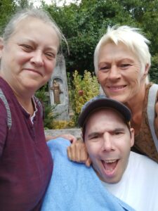 Vor einer kleinen bronzenen Statue posieren zwei Betreuerinnen mit dem lachenden Bewohner Akos für ein gemeinsames Erinnerungsfoto.