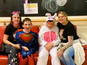 Zwei Bewohner posieren mit zwei Betreuerinnen auf einer roten Couch. Ein Bwohner ist als Superman verkleidet, der andre als Hase.