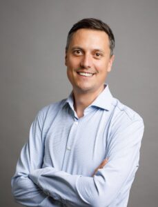 Mario Zagler trägt ein hellblaues Hemd und eine blaue Jeanhose. Seine braunen Haare trägt er kurz. Er lächelt für das Foto freundlich in die Kamera.